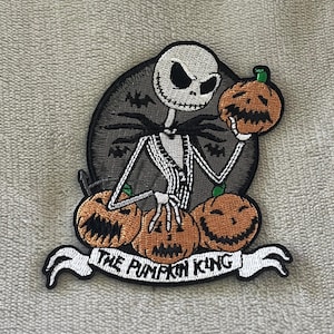 以下が含まれることがあります： ナイトメアー・ビフォア・クリスマスのジャック・スケリントンが描かれた刺繍パッチ。パッチには、カボチャを持ったジャック、下に3つのカボチャ、バナーに「THE PUMPKIN KING」の文字が描かれています。