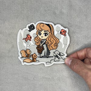 May include: An embroidered patch featuring a cartoon character with long wavy blonde hair wearing a grey dress and holding a brown basket. The character is surrounded by small embroidered animals including squirrels, birds, and a rabbit in a forest setting.