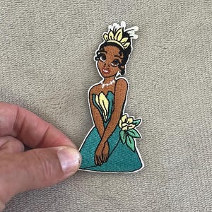 May include: Embroidered patch featuring a cartoon princess in a teal dress with a yellow flower and crown. The princess has brown skin, black hair, and a pearl necklace. The patch is white-edged and appears to be iron-on.