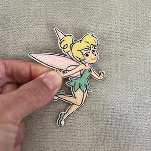 May include: Embroidered patch featuring a cartoon fairy with blonde hair, a green dress, and pink wings. The fairy is in a flying pose with a smiling expression. The patch has a white border and is held by a hand.