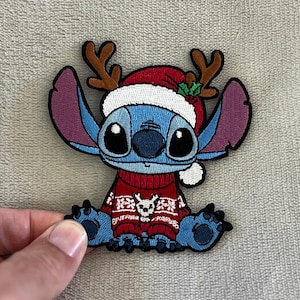 以下が含まれることがあります： リロ＆スティッチのキャラクター、スティッチが赤いクリスマスセーターとサンタ帽、トナカイの角を身につけた青い刺繍パッチ。パッチは黒いアウトラインと様々な色で詳細に描かれています。