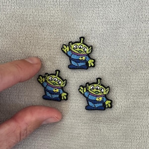 May include: Three green alien embroidered patches with three eyes, a yellow star on their chest, and blue and black clothing.