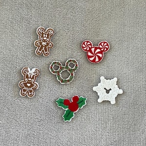 May include: A collection of Christmas-themed embroidered patches. Designs include gingerbread men, Mickey Mouse heads with candy cane and wreath details, a snowflake, and holly. The patches are in shades of brown, red, green, and white.