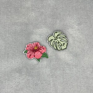 Könnte beinhalten: Zwei gestickte Aufnäher, bestehend aus einer leuchtend rosa Hibiskusblüte mit grünen Blättern und einem hellgrünen Monstera-Blatt. Der Hibiskus hat ein gelbes Zentrum und detaillierte Stickereien, während das Monstera-Blatt deutliche Einschnitte aufweist.