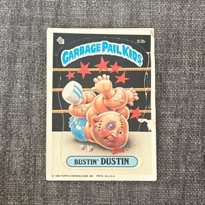 Op de afbeelding: Een vintage Garbage Pail Kids-verzamelkaart met "Bustin' Dustin". De kaart toont een cartoonillustratie van een personage met een blauw oog, een verband en een bloedneus. De titel van de kaart staat in een blauwe doos.