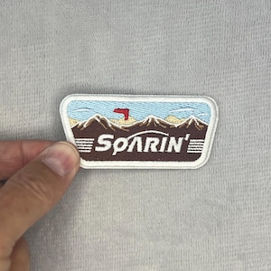 Op de afbeelding: Een rechthoekige geborduurde patch met een witte rand met een berglandschap. Het ontwerp bevat bruine bergen, een lichtblauwe lucht, een klein rood vliegtuigje en de witte tekst "SOARIN'" gecentreerd over de bruine ondergrond.