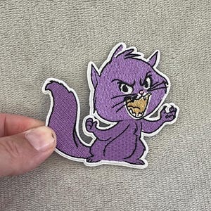 Op de afbeelding: Een paarse geborduurde patch met een boze cartoonkat met een witte rand. De kat laat zijn tanden zien en bevindt zich in een dreigende houding. De patch is bedoeld voor kleding of accessoires.