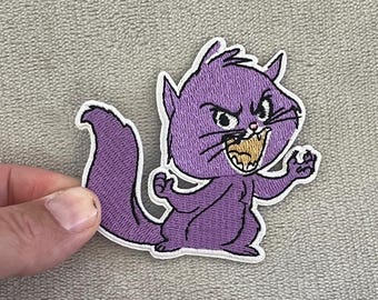 May include: A purple embroidered patch featuring an angry cartoon cat with a white outline. The cat has a wide open mouth, showing its teeth, and is in a threatening pose. The patch is likely for clothing or accessories.