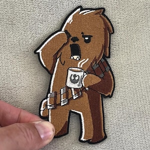 May include: Embroidered patch of Chewbacca from Star Wars, yawning and holding a coffee mug with a Star Wars logo. The patch has a white border and detailed stitching. The character is brown.