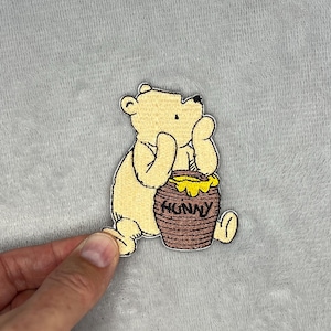 Puede incluir: Parche bordado con un oso amarillo sosteniendo un tarro de miel marrón con la palabra "HUNNY". El oso está sentado y mirando el tarro de miel. El parche está sobre un fondo gris.