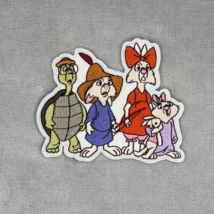 Puede incluir: Un parche bordado que muestra un grupo de cuatro personajes de dibujos animados: una tortuga verde con sombrero marrón, un conejo con túnica azul y sombrero marrón con pluma roja, un conejo con vestido naranja y lazo rojo, y un conejo pequeño con una muñeca.