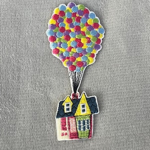Könnte beinhalten: Gestickter Aufnäher mit einem Haus, das von einem Heißluftballon angehoben wird. Der Ballon besteht aus vielen kleinen, farbigen Kreisen. Das Haus hat ein rot, rosa, gelb und blaues Farbschema.