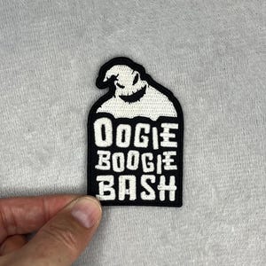 May include: A black and white patch featuring the character Oogie Boogie from The Nightmare Before Christmas. The patch has the words "Oogie Boogie Bash" in white block letters. The patch is rectangular with a curved top.