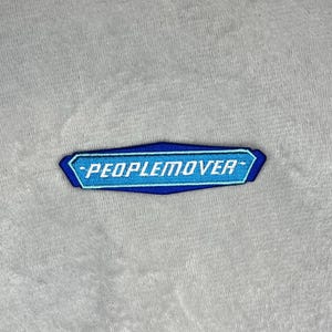 Op de afbeelding: Een blauw en wit geborduurde patch met de tekst "PEOPLEMOVER" in een gestileerd lettertype. De patch heeft een donkerblauwe rand en is geplaatst tegen een lichtgrijze achtergrond.