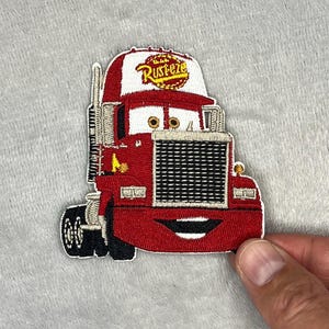 May include: Embroidered patch of a red cartoon truck with a smiling face. The truck has a white and red cap with the word "Rusteze" in gold lettering. The patch is on a grey background.