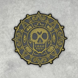 May include: A circular patch with a gold skull design at the centre, surrounded by intricate gold patterns on a grey background. The patch has a black border and a textured appearance, suitable for application on clothing or accessories.