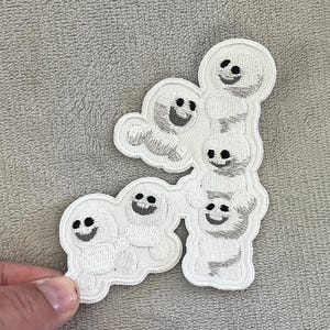 May include: Embroidered white patch featuring a stack of smiling ghost-like figures. Each figure has a simple black smiling face. The patch is on a gray background.