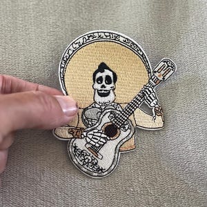 Könnte beinhalten: Gestickter Aufnäher mit einem Skelett-Mariachi-Musiker mit einer Gitarre. Der Aufnäher hat einen beigefarbenen Hintergrund mit einem kreisförmigen Design hinter dem Kopf des Skeletts. Die Gitarre ist weiß mit schwarzen Details.