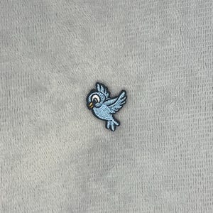 May include: An embroidered patch featuring a small, light blue bird in flight. The bird has a white eye with a black pupil, a small yellow beak, and dark blue outlines defining its wings, body, and tail feathers against a textured grey surface.