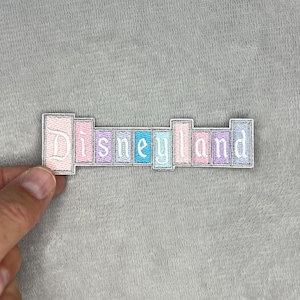 May include: Embroidered patch with the word "Disneyland" in pastel colors. The patch features a white border and is held against a gray fabric background. The letters are in a stylized font.