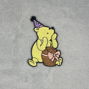 Puede incluir: Un parche bordado que muestra un oso amarillo con un gorro de fiesta morado con un pompón negro. El oso está sentado y apoyado contra un tarro de miel marrón decorado con un lazo rosa claro, todo delineado con hilo de color negro.