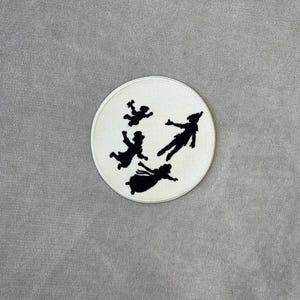 May include: A circular white patch featuring a black silhouette design of four figures in flight. The patch is embroidered with a detailed outline of the characters, set against a soft, gray background. The design evokes a sense of fantasy and adventure.