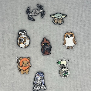 Peut inclure: Un ensemble de huit écussons brodés représentant divers personnages et véhicules de science-fiction, incluant un chasseur TIE, un extraterrestre vert, un droïde rond blanc et orange, une silhouette encapuchonnée, une créature brune, un personnage poilu avec une lance, un droïde bleu et blanc, et un petit véhicule vert et blanc.