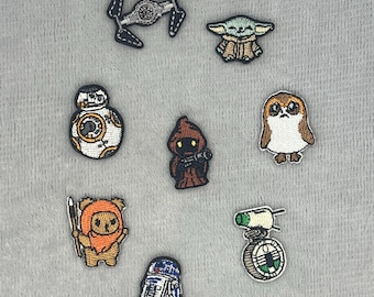 May include: A collection of eight embroidered patches featuring various science fiction characters and vehicles, including a TIE fighter, a green alien, a round white and orange droid, a hooded figure, a brown owl-like creature, a furry creature with a spear, a blue and white droid, and a small green and white vehicle.