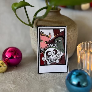 May include: An embroidered iron-on patch featuring a photo booth style image of three characters from the animated film 'The Nightmare Before Christmas'. The characters are Jack Skellington, Sally, and Oogie Boogie. The patch is white with black stitching and a red background.