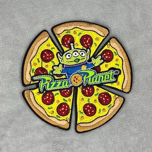 Puede incluir: Parche bordado con un diseño de pizza con pepperoni y un personaje alienígena verde. El parche tiene las palabras "Pizza Planet" en una fuente estilizada. La pizza está dividida en cinco porciones.