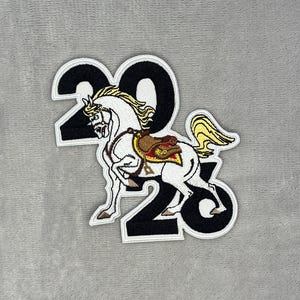 May include: Embroidered patch featuring a white horse with a yellow mane and tail, wearing a saddle. The horse is positioned within the numbers "2023" in black with a white border. The patch is on a gray background.