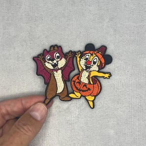 Peut inclure: Un écusson brodé représentant deux tamias de dessin animé en costumes d'Halloween. L'un porte un costume de chauve-souris violet avec des ailes, l'autre un costume de citrouille orange et un serre-tête à oreilles de souris, tous deux souriants.