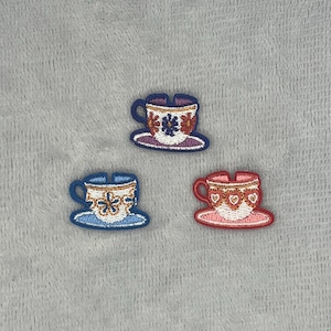 Puede incluir: Tres parches bordados con forma de tazas de té sobre platillos. El parche superior presenta una taza morada y blanca con un diseño floral. El parche inferior izquierdo es azul y blanco con un patrón de flores. El inferior derecho es rojo y blanco con corazones.