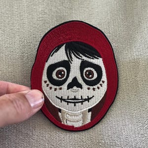 Puede incluir: Parche bordado con un rostro de calavera estilizado con una capucha roja. El parche tiene un borde negro y representa una calavera sonriente con ojos marrones y un diseño detallado. El parche es para ropa o accesorios.