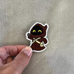 May include: A cartoon patch featuring a Jawa character from Star Wars. The Jawa is depicted in a brown hooded robe with a yellow utility belt and a small pouch. The Jawa has a black face with yellow eyes and a small, round object in its hand.