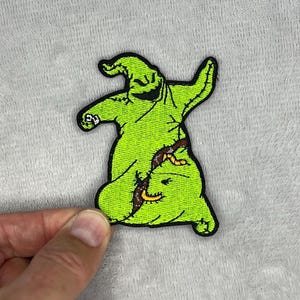 Puede incluir: Un parche bordado verde con un personaje de dibujos animados con un contorno negro. El personaje tiene una cara sonriente y está sentado. El parche está sostenido por una mano sobre un fondo gris.