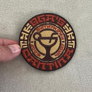 May include: Round patch with a cocktail glass design and text in brown, tan, and red. The patch is held by a hand. The design is circular.