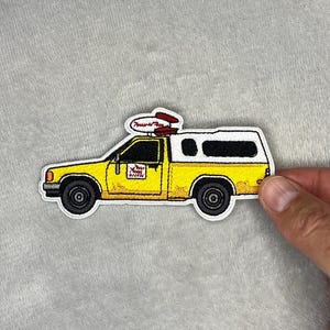 Puede incluir: Parche bordado con una camioneta amarilla con una capota blanca. La camioneta tiene ruedas negras y una tabla de surf roja en la parte superior. El parche incluye el texto "Pizza Planet" en el lateral.