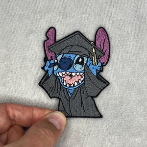 May include: Embroidered patch featuring the character Stitch from Lilo & Stitch, wearing a graduation cap and gown. The patch is blue, black, and pink, with a gold tassel on the cap. The design is detailed and colorful.