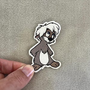 May include: Embroidered patch featuring a cartoon lemur character with white fur, brown body, and blue eyes. The patch has a white border and is held by a hand against a neutral background. The lemur is in a seated position.