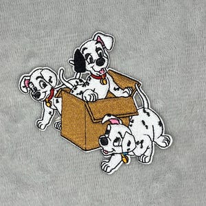 Puede incluir: Parche bordado con tres cachorros dálmatas. Dos cachorros están fuera de una caja de cartón marrón, y uno está dentro. Los cachorros tienen manchas negras, collares rojos y etiquetas doradas. El parche está sobre un fondo de tela gris.