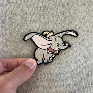 Könnte beinhalten: Gestickter Aufnäher mit Dumbo, dem fliegenden Elefanten. Der Aufnäher hat einen grauen Körper, große, rosa gefütterte Ohren und einen gelben Hut. Schwarz umrandet, ist der Aufnäher ein skurriles Accessoire für Kleidung oder Taschen.