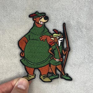 Peut inclure: Écusson brodé représentant un ours et un renard en tenues vertes. L'ours porte un chapeau et le renard tient un arc. L'écusson est posé sur un fond gris clair.