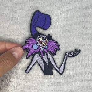 May include: An embroidered patch featuring a cartoon character with pale skin, a wide grin, and yellow eyes. The character wears a tall, curved purple hat, a dark purple feathered collar, and light blue circular earrings against a black outfit.