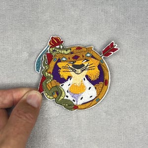 May include: An embroidered patch featuring a cartoon lion wearing a crown and a white ermine-trimmed cape. A green snake with a small red hat is coiled around the lion, and a red-feathered arrow is positioned behind the circular gold frame.