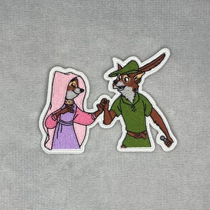 Peut inclure: Un écusson brodé représentant deux renards anthropomorphes se tenant la main. La figure de gauche porte une cape à capuche rose sur une robe violette. La figure de droite porte une tunique verte, une ceinture marron et un chapeau vert avec une plume marron.