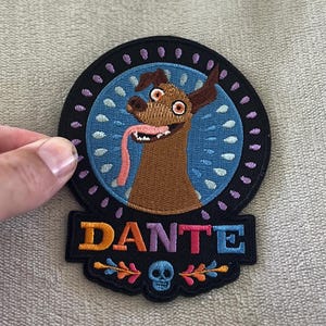 Könnte beinhalten: Gestickter Aufnäher mit Dante, einem braunen Hund mit langer rosa Zunge, aus dem Film Coco. Der Aufnäher hat einen schwarzen Rand mit dem Namen "DANTE" in bunten Buchstaben und einem blauen Totenkopf.