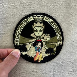 May include: Round embroidered patch featuring a cartoon image of Snow White and the Evil Queen. The Evil Queen is holding an apple, and Snow White is wearing a yellow dress. The patch has a black background with green and gold accents.