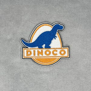 May include: An embroidered patch featuring a blue dinosaur silhouette centered within an oval frame. The background of the oval is white and orange. Below the dinosaur, a rectangular orange banner contains the white text DINOCO in capital letters.
