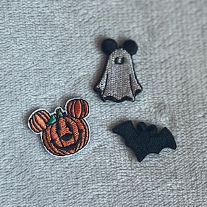 Könnte beinhalten: Drei Halloween-Stick-Patches. Ein Kürbis mit Micky-Maus-Ohren, ein Geist mit Micky-Maus-Ohren und eine schwarze Fledermaus. Die Patches befinden sich auf einer grauen, strukturierten Oberfläche.